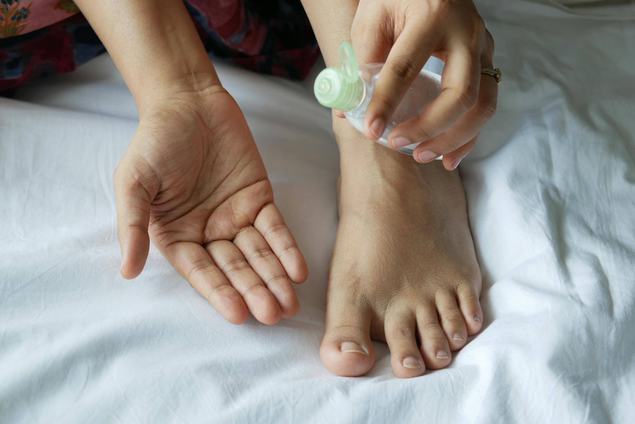 Close-up of hands and foot using a plastic bottle of oil on a bed. Skincare routine concept.